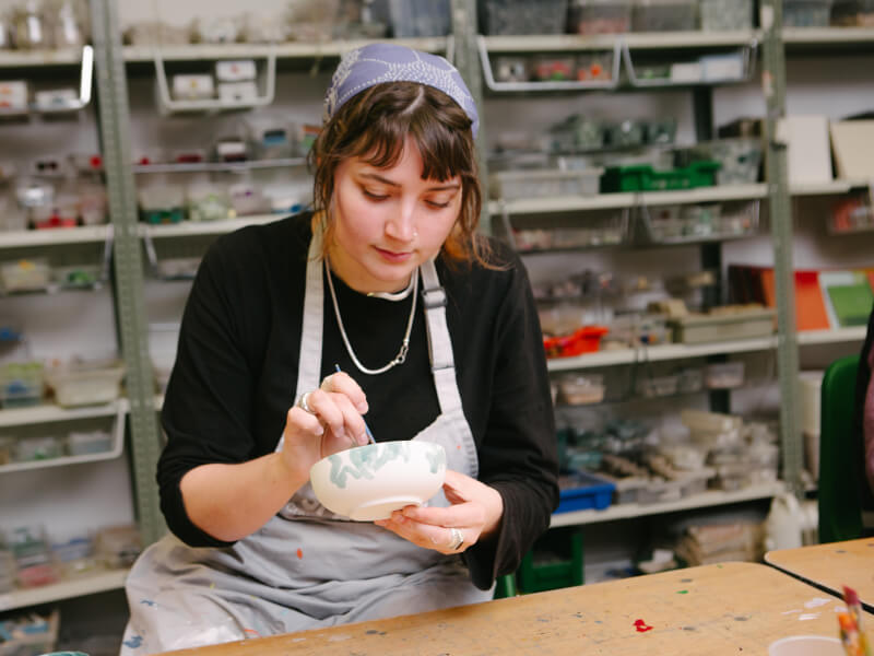 A woman is sitting at a table, painting a green design onto a plain white pottery bowl